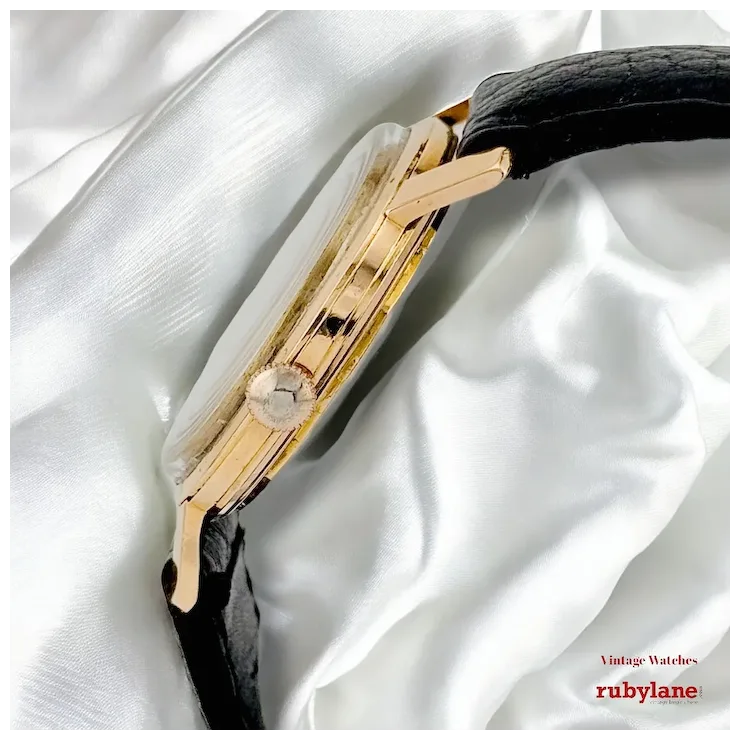 Gold vintage watch with black strap on a white fabric background, featuring the Ruby Lane brand.