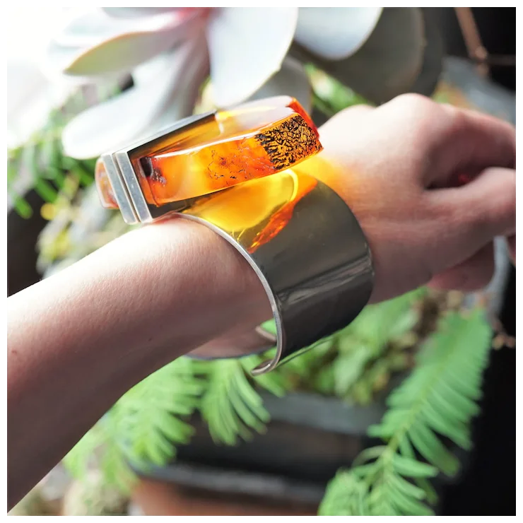 Person wearing a futuristic bracelet with a glowing orange center against a blurred green plant background