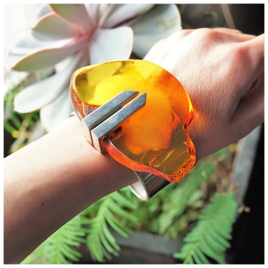 Hand wearing a large orange and silver bracelet with plants in the background