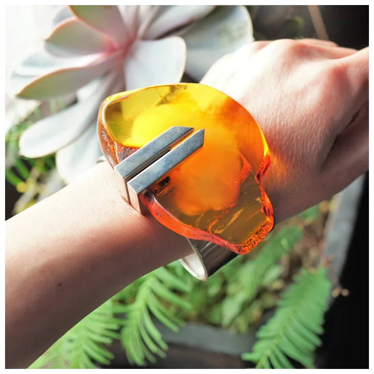 Hand wearing a large orange and silver bracelet with plants in the background
