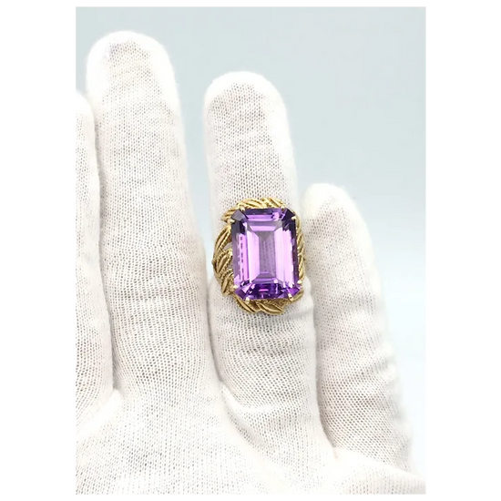Gold ring with a purple gemstone held by a white gloved hand on a light gray background