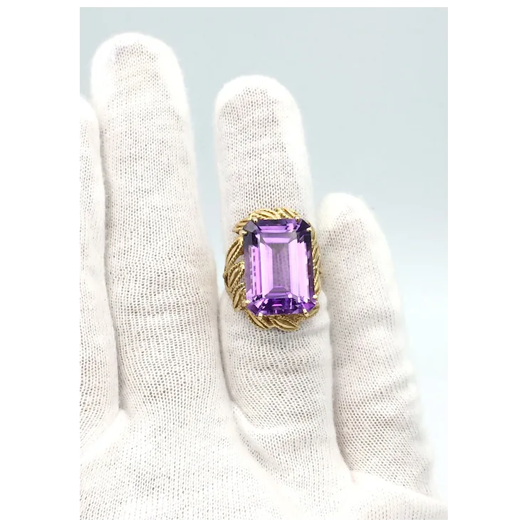 Gold ring with a purple gemstone held by a white gloved hand on a light gray background