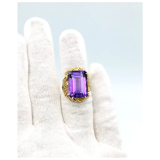 Ring with a purple gemstone held by a white gloved hand on a light blue background