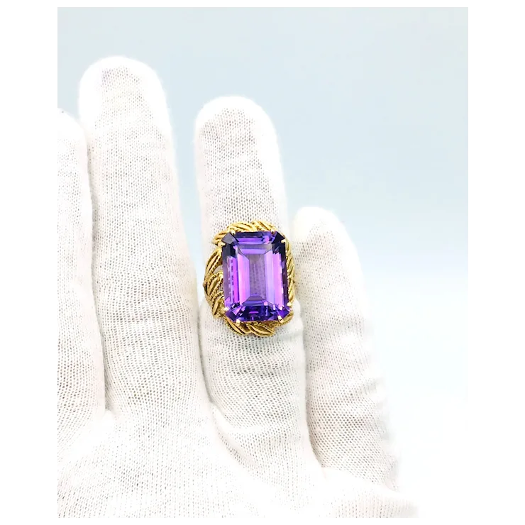 Ring with a purple gemstone held by a white gloved hand on a light blue background