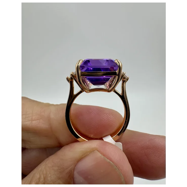 Gold ring with a purple gemstone held between fingers against a neutral background