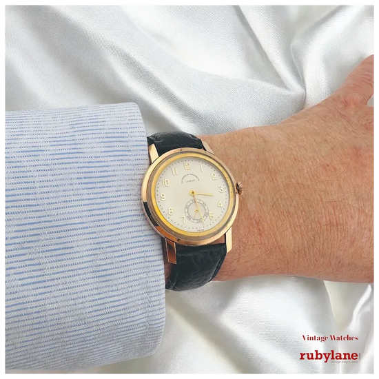 Gold watch with black strap on a wrist against a white fabric background, featuring the Rubylane brand.
