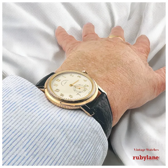 Hand wearing a vintage watch with a white background, featuring Rubylane branding.