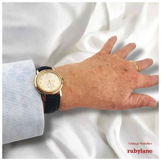 Hand wearing a gold and black watch on a white fabric background with Rubylane branding.
