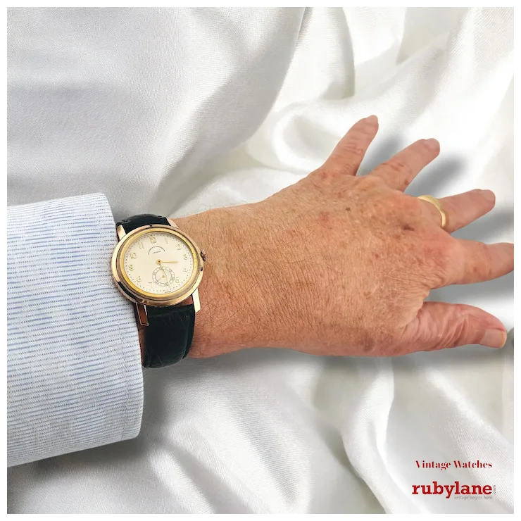 Hand wearing a gold and black watch on a white fabric background with Rubylane branding.