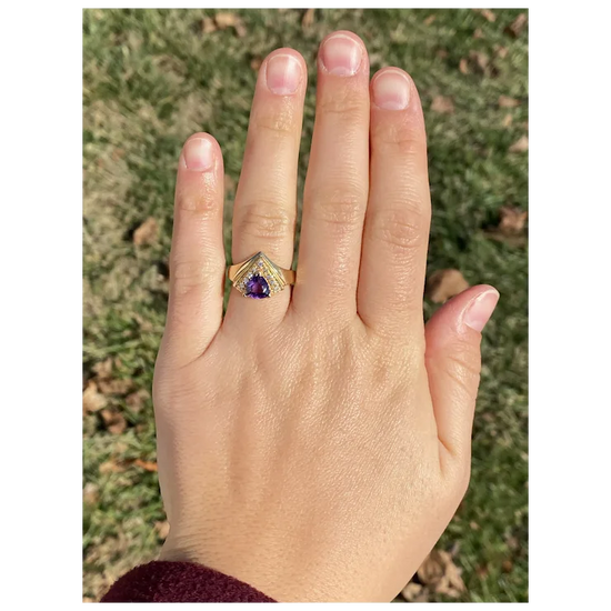 Hand wearing a gold ring with a purple gemstone against a grassy background