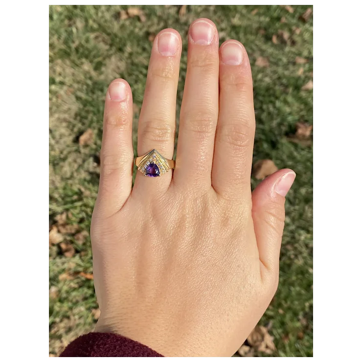 Hand wearing a gold ring with a purple gemstone against a grassy background