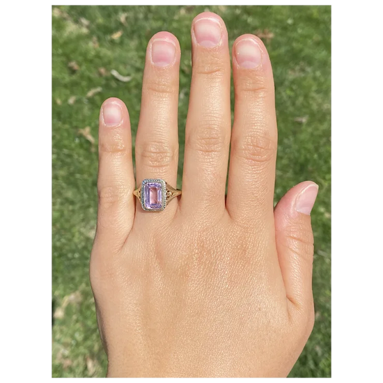 Hand wearing a ring with a purple gemstone against a grassy background