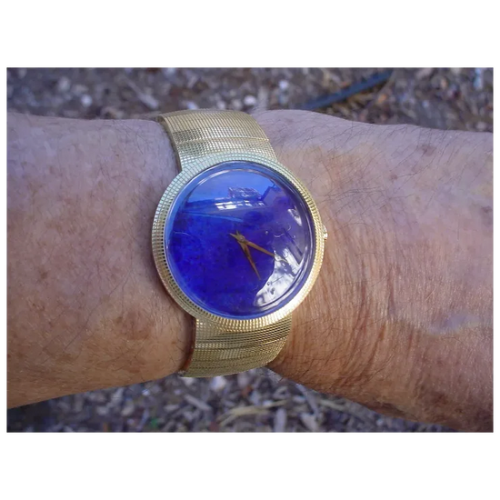 Gold wristwatch with a blue face on a wrist, blurred natural background