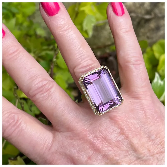 Hand wearing a ring with a purple gemstone against a blurred green background