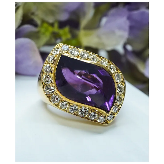 Gold ring with a purple gemstone on a white surface with blurred flowers in the background